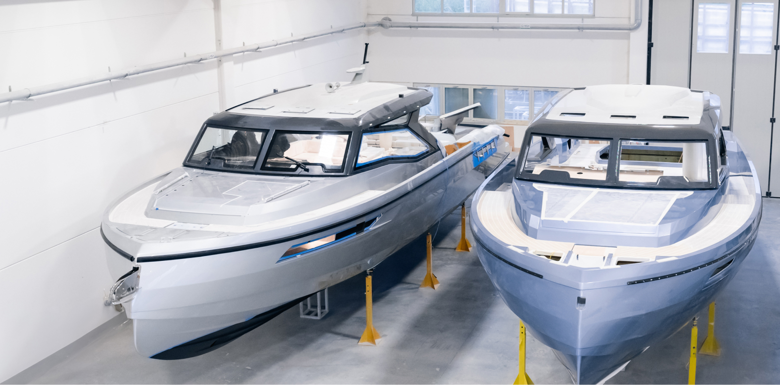 Two luxury motor yachts stored indoors on yellow stands, with white and blue hulls side by side in a workshop.