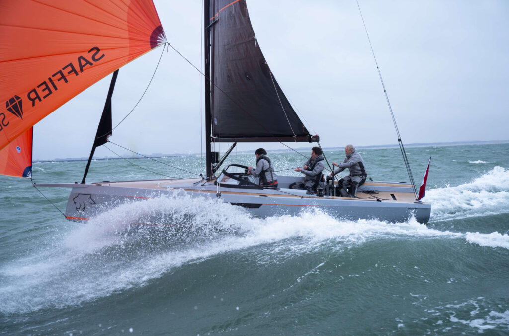 Sleek racing sailboat with an orange spinnaker on the left, three sailors maneuvering as waves splash.
