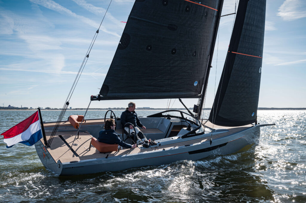 Sailboat on open water with two sailors on a wooden deck; black sails and a Dutch flag at the stern.