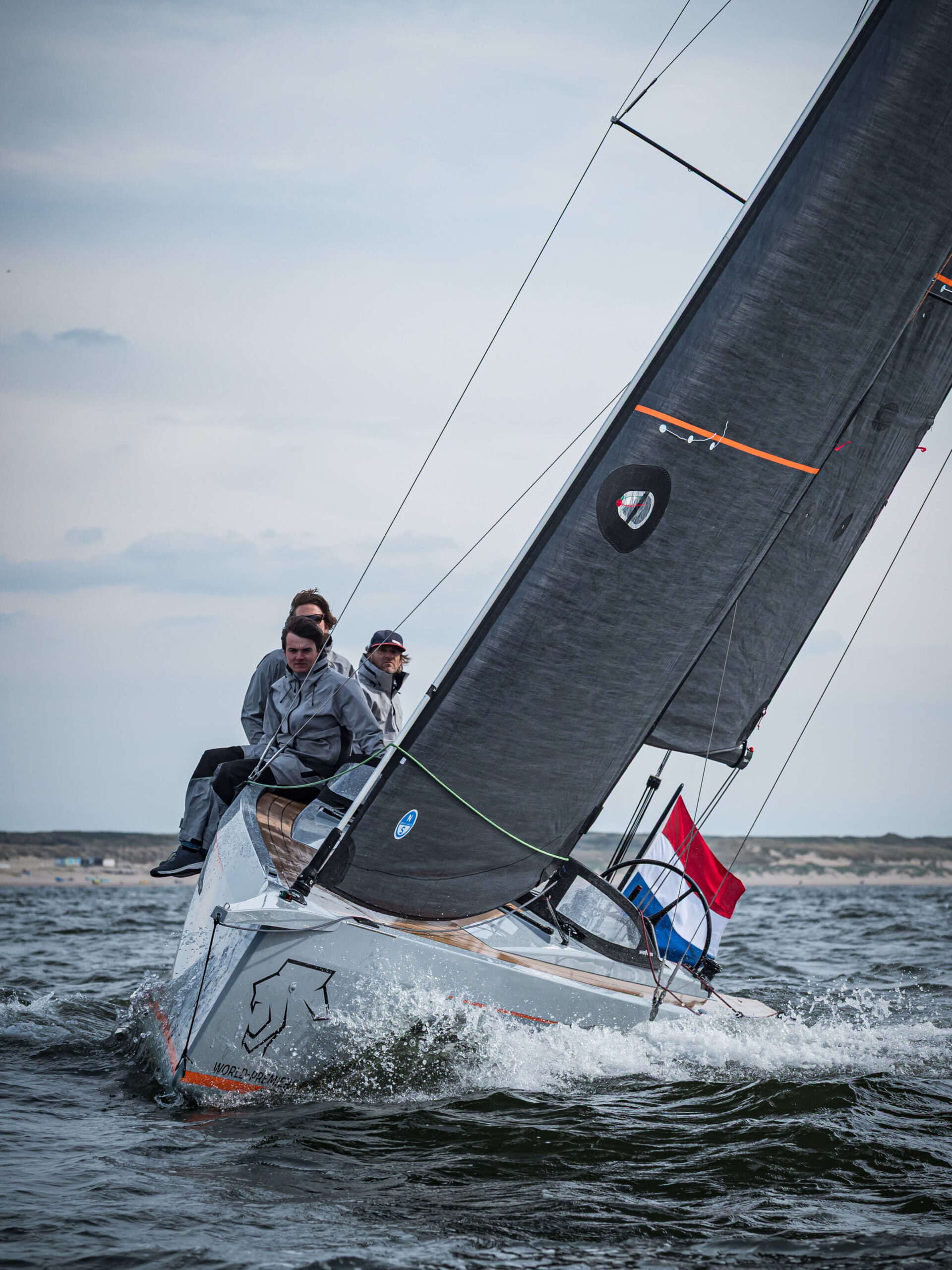 Sailing team on a racing sailboat heeling to the left, cutting through waves near the coastline with passengers on deck, wind in the sails.