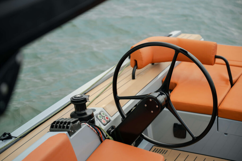 Boat cockpit with a black steering wheel and orange cushioned seats, water in the background, view from the side.